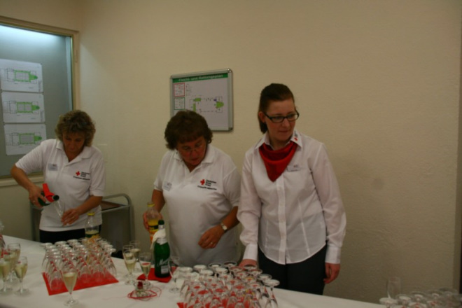Drei Damen stehen hinter einem Tisch und gießen Sekt aus Flaschen in Sektgläser.