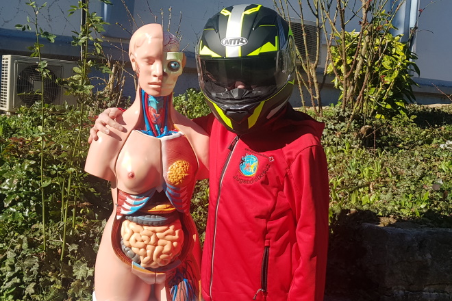Ein Jugendlicher mit einem Motoradhelm hält eine Puppe mit eintnehmbaren Orgenen im Arm. Motorradhelm und die Puppe sind mit dem Sozial-Coin-Geld angeschafft worden.