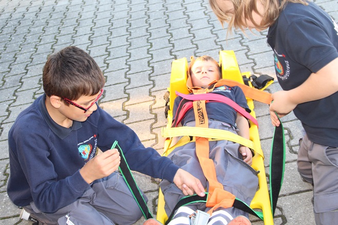 Zwei Jugendliche sichern einen Patient mit Gurten auf einem Spineboard, um ihn für den Transport vorzubereiten.